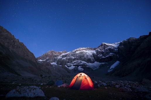 A brightly lit tent set against a backdrop of rugged, snow-capped mountains under a clear, starry night sky.