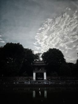 A serene landscape featuring a traditional pavilion structure with a tiled roof situated in front of a backdrop of lush, dense trees. The foreground includes a reflective body of water that mirrors the pavilion's image, creating a symmetrical and tranquil scene. Overhead, dramatic clouds stretch across a moody sky, adding depth and contrast to the composition.