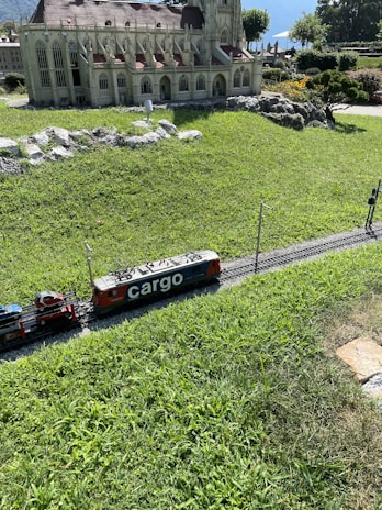 A model train labeled 'cargo' travels on miniature tracks next to a detailed architectural model of a building with gothic-style windows and a sloped roof. The scene is set amidst well-manicured grass and surrounded by small rocks and shrubs, with trees and a scenic backdrop in the distance.