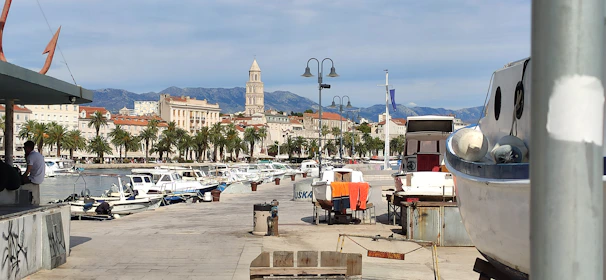 A picturesque marina with numerous small boats docked alongside a stone walkway. Palm trees line the opposite side of the waterfront, with historic buildings and a tall bell tower visible in the background. A person is standing near a small kiosk, and there's a large boat on land in the foreground.