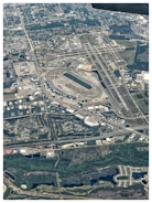 An aerial view of an airport with multiple runways and taxiways. Surrounding the airport, there are numerous buildings, roads, and a racetrack. The area is integrated with greenery, waterways, and urban structures. Residential and industrial zones are visible, with various streets and infrastructures.
