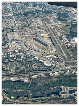 An aerial view of an airport with multiple runways and taxiways. Surrounding the airport, there are numerous buildings, roads, and a racetrack. The area is integrated with greenery, waterways, and urban structures. Residential and industrial zones are visible, with various streets and infrastructures.