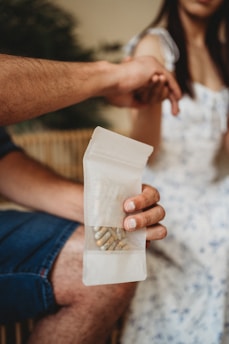 A person holds a transparent bag containing several capsules while another person sits nearby. The scene appears to be indoors, and part of the focus is on the interaction between the two individuals.