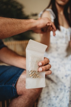 A person holds a transparent bag containing several capsules while another person sits nearby. The scene appears to be indoors, and part of the focus is on the interaction between the two individuals.