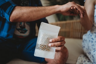 A person holds a transparent bag containing capsules or pills. Another hand is reaching out towards the person holding the bag. The setting appears relaxed, possibly in an indoor environment with a wicker or wooden element in the background.