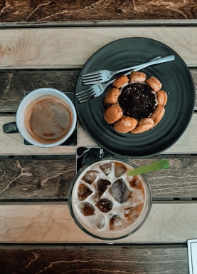 A table setting featuring a dark plate with a dessert encircled by small cookies or pastries, accompanied by a fork. Next to it, a cup of coffee with light brown foam and a glass of iced coffee with a green straw. The table has a rustic, wooden texture.