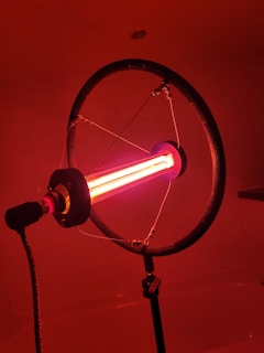 A lamp with a ring-shaped structure surrounding a bright red fluorescent tube, mounted on a stand in a dimly lit room. The lighting casts a warm, reddish glow throughout the space.