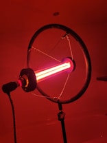 A lamp with a ring-shaped structure surrounding a bright red fluorescent tube, mounted on a stand in a dimly lit room. The lighting casts a warm, reddish glow throughout the space.