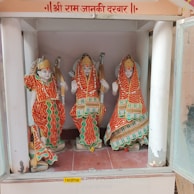 Three intricately designed statues are dressed in colorful traditional Indian attire, holding bows, and seated within a shrine. The garments feature red, orange, and green colors with elaborate patterns and embellishments. The backdrop suggests a sacred setting.