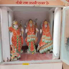 Three intricately designed statues are dressed in colorful traditional Indian attire, holding bows, and seated within a shrine. The garments feature red, orange, and green colors with elaborate patterns and embellishments. The backdrop suggests a sacred setting.
