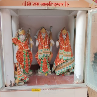 Three intricately designed statues are dressed in colorful traditional Indian attire, holding bows, and seated within a shrine. The garments feature red, orange, and green colors with elaborate patterns and embellishments. The backdrop suggests a sacred setting.