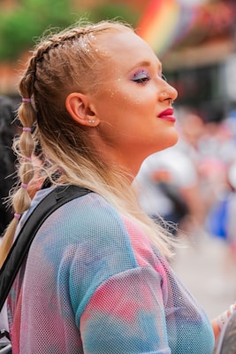 A person with braided hair adorned with glitter makeup stands in a vibrant outdoor setting. They wear a colorful shirt with pastel tones and gaze contentedly into the distance. In the background, a rainbow flag is visible, adding to the festive and inclusive atmosphere.