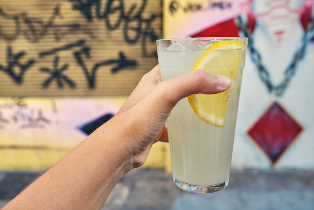 a hand holding a glass of lemonade in front of a graffiti covered wall