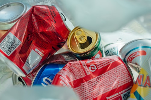 A collection of crushed and discarded beverage cans with various labels and colors, including red, blue, and green. The cans appear to be in a recycling or waste setting.