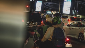 Several motorcyclists and cars are stuck in traffic at night. Bright lights from billboards and the vehicles' taillights illuminate the scene. A person wearing a green and black helmet is seen from behind, seated on a motorcycle, wearing a backpack.
