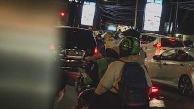 Several motorcyclists and cars are stuck in traffic at night. Bright lights from billboards and the vehicles' taillights illuminate the scene. A person wearing a green and black helmet is seen from behind, seated on a motorcycle, wearing a backpack.