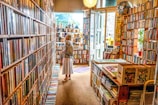 A person is standing in a cozy bookstore or library, surrounded by shelves filled with books. The interior has a warm, inviting atmosphere, with wooden furniture and an open door letting in natural light. Comics and various types of books are organized across the shelves.