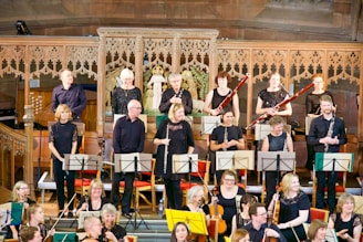 A group of musicians are gathered on a stage with ornate wooden carvings in the background. They are holding various orchestral instruments like clarinets, bassoons, and violins, and appear to be preparing for a performance. The musicians are dressed in black attire.