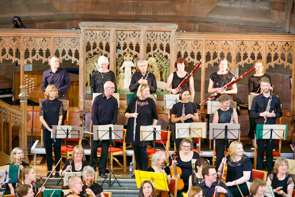 A group of musicians are gathered on a stage with ornate wooden carvings in the background. They are holding various orchestral instruments like clarinets, bassoons, and violins, and appear to be preparing for a performance. The musicians are dressed in black attire.