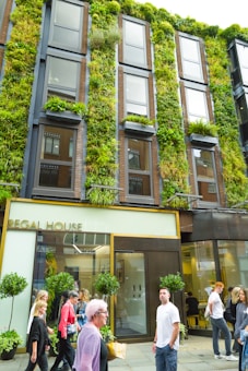 The image depicts a multi-story building with a vertical garden covering its facade, featuring various types of greenery and plants. Several people are walking on the sidewalk in front of the building, and the ground floor has a sign that reads 'Regal House'. The windows are large and the structure is modern, blending nature with urban architecture.