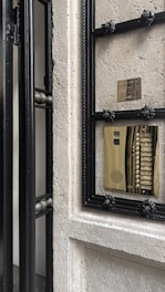 An ornate door with a black metallic frame, partially opened, reveals a brass intercom system embedded in the wall. The stone wall appears textured and there is a small plaque above the intercom.
