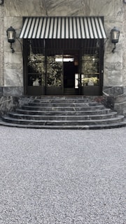 A large window with a striped awning above a set of stone steps. The steps are made of a polished, dark marble leading to a glass door. Two vintage-style lanterns flank the doorway. The ground is covered with small grey pebbles.