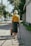 An elderly woman walks down a sunlit sidewalk with a cane in one hand and a bag in the other. She wears a yellow top and a gray skirt, and the background features trees and houses along the street.