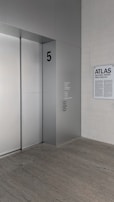 A modern, minimalist interior with an elevator and a wall-mounted plaque. The elevator door is sleek and metallic, marked with the number '5' in bold. Nearby, a sign displays the words 'ATLAS', 'MICHAEL HEIZER', and 'PINO PASCALI' in large text.