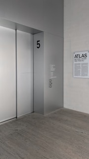 A modern, minimalist interior with an elevator and a wall-mounted plaque. The elevator door is sleek and metallic, marked with the number '5' in bold. Nearby, a sign displays the words 'ATLAS', 'MICHAEL HEIZER', and 'PINO PASCALI' in large text.