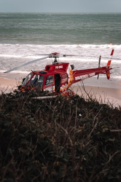 A red rescue helicopter is parked on a sandy beach near the ocean. The helicopter bears markings indicating it is a rescue vehicle. The background shows waves crashing on the shore, and there is a glimpse of greenish vegetation in the foreground.