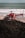 A red rescue helicopter is parked on a sandy beach near the ocean. The helicopter bears markings indicating it is a rescue vehicle. The background shows waves crashing on the shore, and there is a glimpse of greenish vegetation in the foreground.