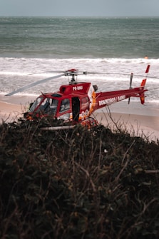 A red rescue helicopter is parked on a sandy beach near the ocean. The helicopter bears markings indicating it is a rescue vehicle. The background shows waves crashing on the shore, and there is a glimpse of greenish vegetation in the foreground.