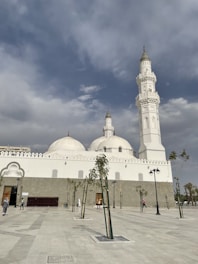 A large white mosque with distinctive domes and minarets under a partly cloudy sky. The mosque has intricate architectural details on its facade. Sparse trees and lampposts line the courtyard, and a few people are walking or standing near the entrance.