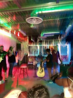 A small music performance venue featuring a band setup. Various musical instruments such as a drum set and guitar are visible. The room is dimly lit with colorful stage lights creating a vibrant and energetic atmosphere. Several people are present, engaging with the performance.