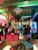 A small music performance venue featuring a band setup. Various musical instruments such as a drum set and guitar are visible. The room is dimly lit with colorful stage lights creating a vibrant and energetic atmosphere. Several people are present, engaging with the performance.