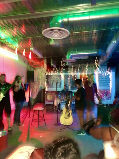 A small music performance venue featuring a band setup. Various musical instruments such as a drum set and guitar are visible. The room is dimly lit with colorful stage lights creating a vibrant and energetic atmosphere. Several people are present, engaging with the performance.