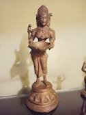 A bronze-colored statue of a deity holding a bowl, with intricate patterns on the clothing and a serene expression on the face. The statue stands on a detailed pedestal, and the entire figure has a polished, metallic appearance.
