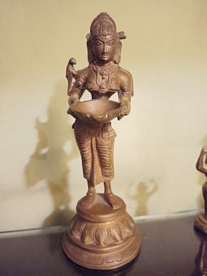 A bronze-colored statue of a deity holding a bowl, with intricate patterns on the clothing and a serene expression on the face. The statue stands on a detailed pedestal, and the entire figure has a polished, metallic appearance.