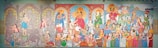 A vibrant mural with three main sections depicting various scenes, including religious and cultural rituals. Central focus on a deity surrounded by worshippers, musicians, and people in traditional attire engaging in celebrations. The left section has muted colors with intricate patterns and figures, while the middle and right sections are more colorful with detailed depictions of festivities and community gatherings.