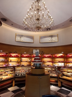 A bakery display with an ornate chandelier hanging from a high ceiling. The interior features warm lighting and wooden shelves filled with colorful packaged goods and various pastries. A tiered cake sits prominently on a round, wooden stand in the center.