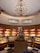 A bakery display with an ornate chandelier hanging from a high ceiling. The interior features warm lighting and wooden shelves filled with colorful packaged goods and various pastries. A tiered cake sits prominently on a round, wooden stand in the center.