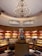A bakery display with an ornate chandelier hanging from a high ceiling. The interior features warm lighting and wooden shelves filled with colorful packaged goods and various pastries. A tiered cake sits prominently on a round, wooden stand in the center.