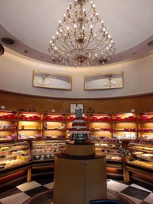 A bakery display with an ornate chandelier hanging from a high ceiling. The interior features warm lighting and wooden shelves filled with colorful packaged goods and various pastries. A tiered cake sits prominently on a round, wooden stand in the center.