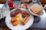 A hearty breakfast spread featuring sunny-side-up eggs, crispy bacon, sausage, hash browns, grilled tomato, and sautéed mushrooms on a plate. Accompanying the meal are a basket of toasted bread, a glass of red juice, a small bowl of fruit salad, and another bowl of baked beans. Cutlery is placed neatly to the side.