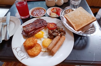 A hearty breakfast spread featuring sunny-side-up eggs, crispy bacon, sausage, hash browns, grilled tomato, and sautéed mushrooms on a plate. Accompanying the meal are a basket of toasted bread, a glass of red juice, a small bowl of fruit salad, and another bowl of baked beans. Cutlery is placed neatly to the side.