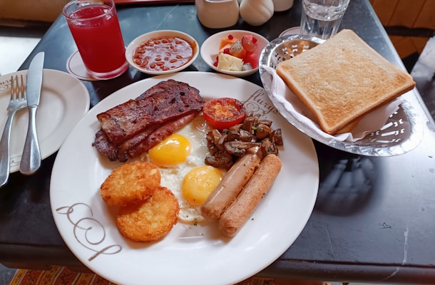 A hearty breakfast spread featuring sunny-side-up eggs, crispy bacon, sausage, hash browns, grilled tomato, and sautéed mushrooms on a plate. Accompanying the meal are a basket of toasted bread, a glass of red juice, a small bowl of fruit salad, and another bowl of baked beans. Cutlery is placed neatly to the side.