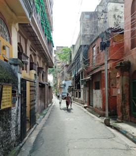 A narrow street lined with old, weathered buildings featuring faded colors and visible wear. Overhead wires crisscross the street, and a few people, including a rickshaw puller, are visible further down the lane. A sign with Bengali script is present on the left side of a building.