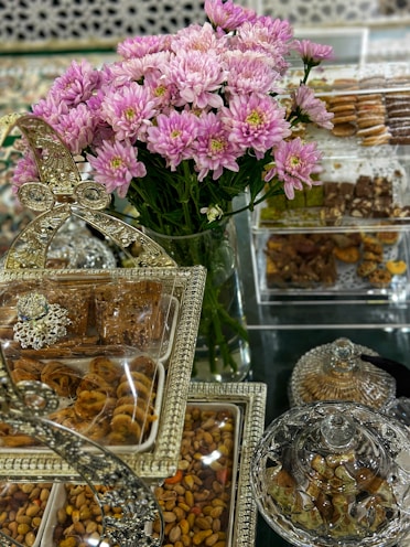 A lavish display featuring a bouquet of pink and white flowers in the center, surrounded by an assortment of sweets and nuts presented in ornate, decorative containers. The setup includes clear glass and metallic elements, giving a luxurious and festive appearance.