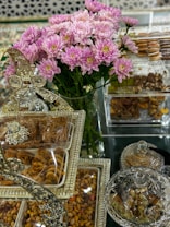 A lavish display featuring a bouquet of pink and white flowers in the center, surrounded by an assortment of sweets and nuts presented in ornate, decorative containers. The setup includes clear glass and metallic elements, giving a luxurious and festive appearance.