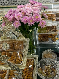 A lavish display featuring a bouquet of pink and white flowers in the center, surrounded by an assortment of sweets and nuts presented in ornate, decorative containers. The setup includes clear glass and metallic elements, giving a luxurious and festive appearance.